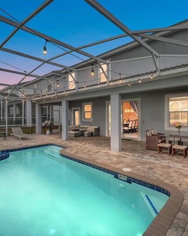 Screened pool deck at dusk with illuminated swimming pool, spa, lounge chairs, and modern house with open doors.