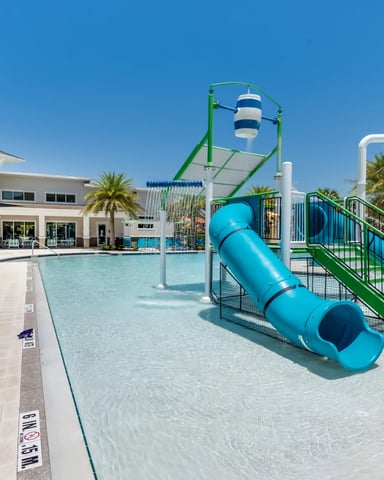 Resort pool with shallow children's area featuring blue water slide, green play structure, and loungers under palm trees