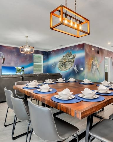 Dining room with wood table, blue place settings, gray chairs, gray sectional sofa, and Star Wars-themed wall murals with spacecraft.