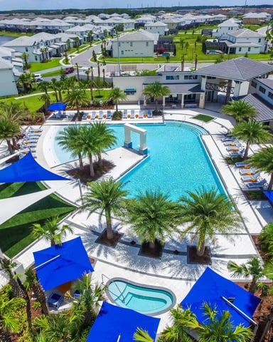 Aerial view of resort community pool with blue shade structures, palm trees, loungers, and surrounding residential homes with waterfront views