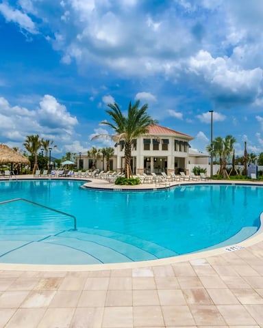Resort pool complex with turquoise water, white clubhouse, palm trees, and thatched-roof cabana under blue sky