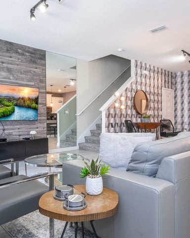 Open-plan living room with gray sectional sofa, wall-mounted TV, wooden accent wall, and staircase with patterned wallpaper.